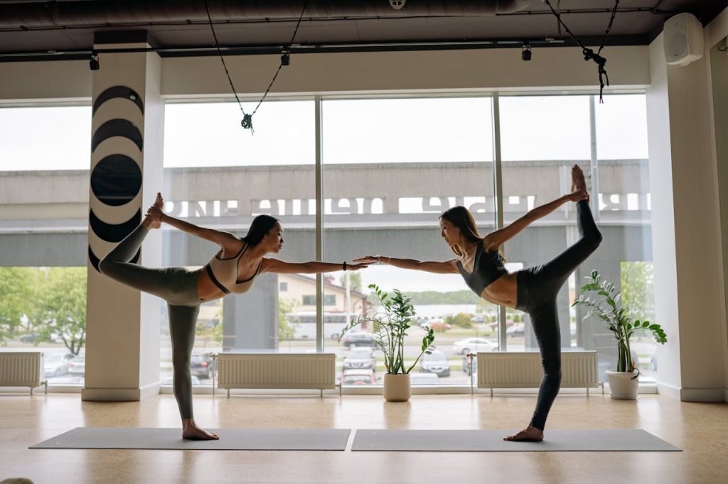 Two women perform a balanced yoga pose in a bright, modern studio, highlighting strength and flexibility.
